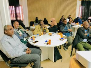 Group of adults seated around round tables in a conference room, listening attentively during a meeting; snacks and beverages on the tables, some attendees in traditional attire.
