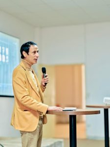 Man in a tan blazer speaks into a handheld microphone at a round podium in an indoor conference room