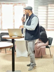 Man wearing a flat cap and vest speaks into a handheld microphone beside a laptop on a round table, with a seated person in the background.