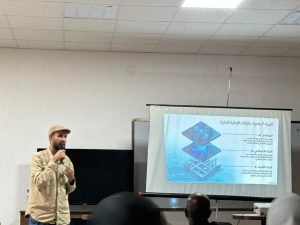 Man in a beige shirt and cap speaks into a microphone at a classroom lectern beside a projector screen displaying a blue multi-layer diagram with Arabic text.