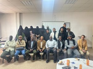 Group portrait of men seated in the front row with women in black niqabs standing behind them in a meeting room. In the foreground is a round table with snacks and drinks.