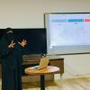 Woman in a black niqab giving a presentation with a laptop on a round table and a projected slide behind her.