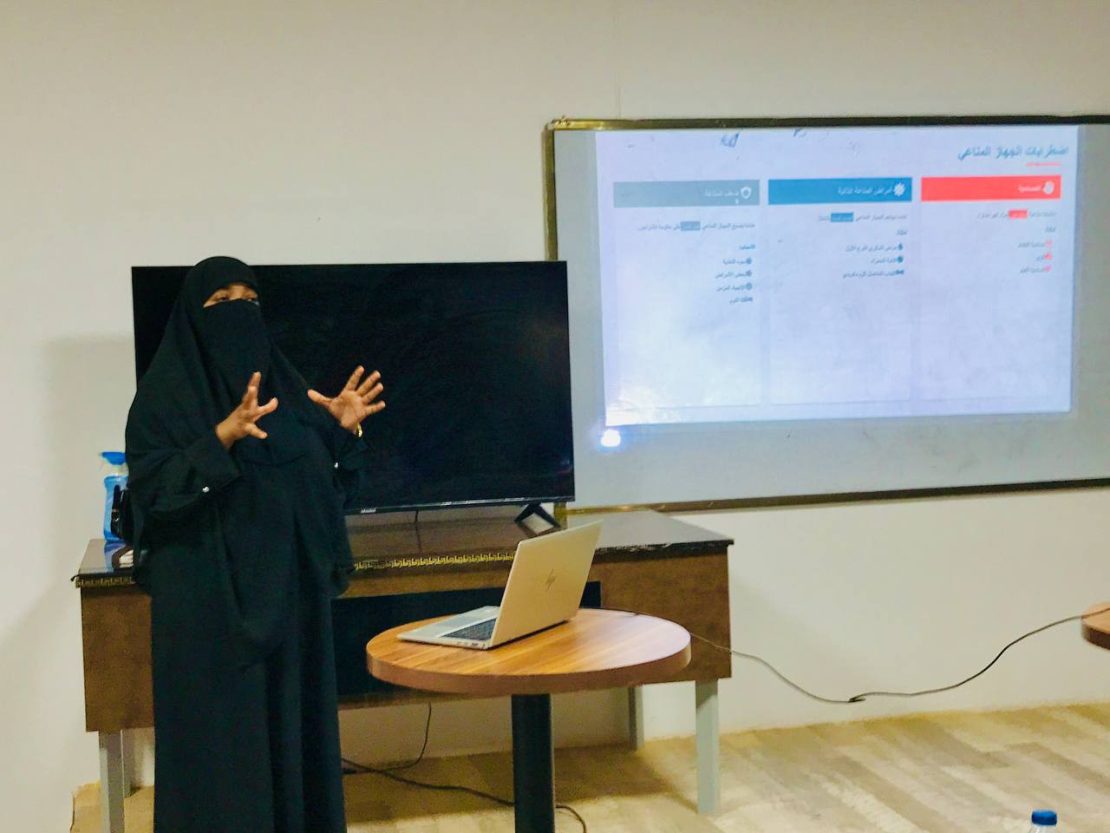Woman in a black niqab giving a presentation with a laptop on a round table and a projected slide behind her.