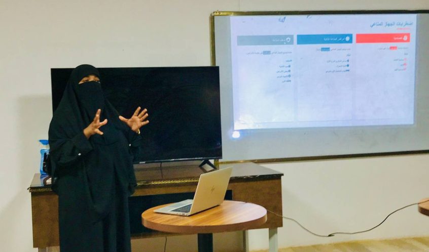 Woman in a black niqab giving a presentation with a laptop on a round table and a projected slide behind her.