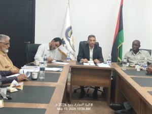 Four men sit around a conference table in a meeting room with flags in the background and papers, bottles, and snacks on the table; timestamp visible at bottom: May 10, 2026 10:36 AM.