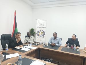 Five men sit around a conference table in a bright room, with a flag and a vertical banner in the background.