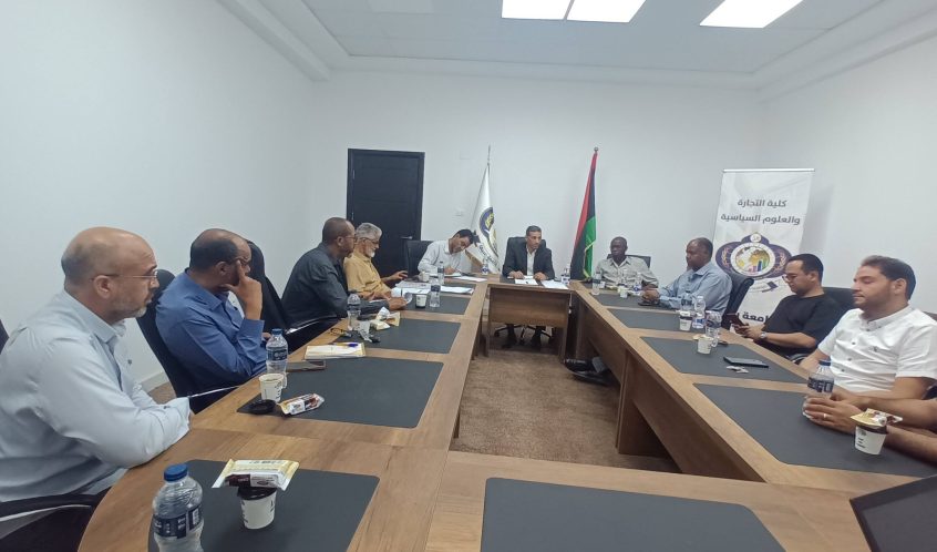 Group of people in a formal meeting around a long conference table with notebooks, water bottles, and snacks; flags and a banner in the background.