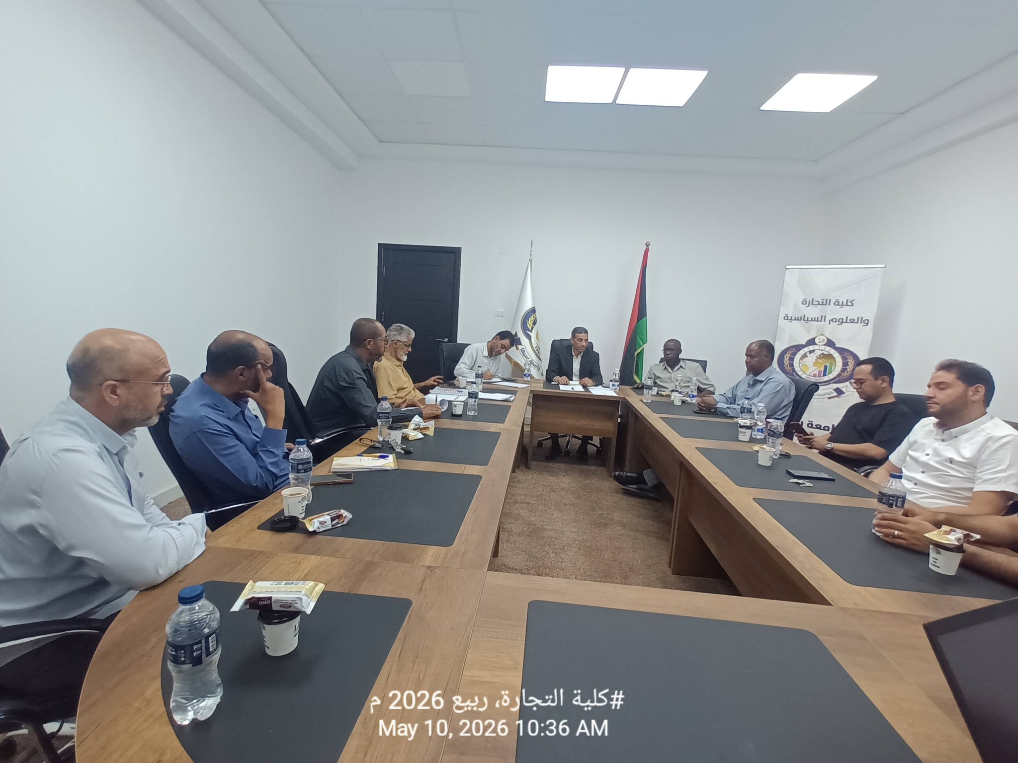 Group of people in a formal meeting around a long conference table with notebooks, water bottles, and snacks; flags and a banner in the background.