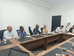 People seated around a large conference table in a meeting room, discussing documents with laptops, coffee cups, and water bottles visible; a flag stands by the wall in the background.