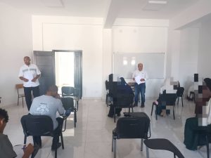 WhatsApp Image 2026-07-06 at 112150 - كلية التجارة والعلوم السياسية Two men stand at the front of a bright classroom presenting, while attendees sit at desks and take notes in a training session with a whiteboard in the background.