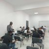 Instructor in a gray suit stands at the front of a bright classroom as students write in notebooks.