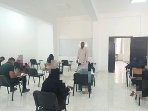 WhatsApp Image 2026-07-06 at 112151 3 - كلية التجارة والعلوم السياسية Instructor standing at the front of a bright classroom near a whiteboard while students sit at desks and take notes. The door is open to the right.