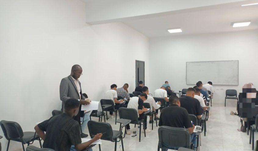 Instructor in a gray suit stands at the front of a bright classroom as students write in notebooks.
