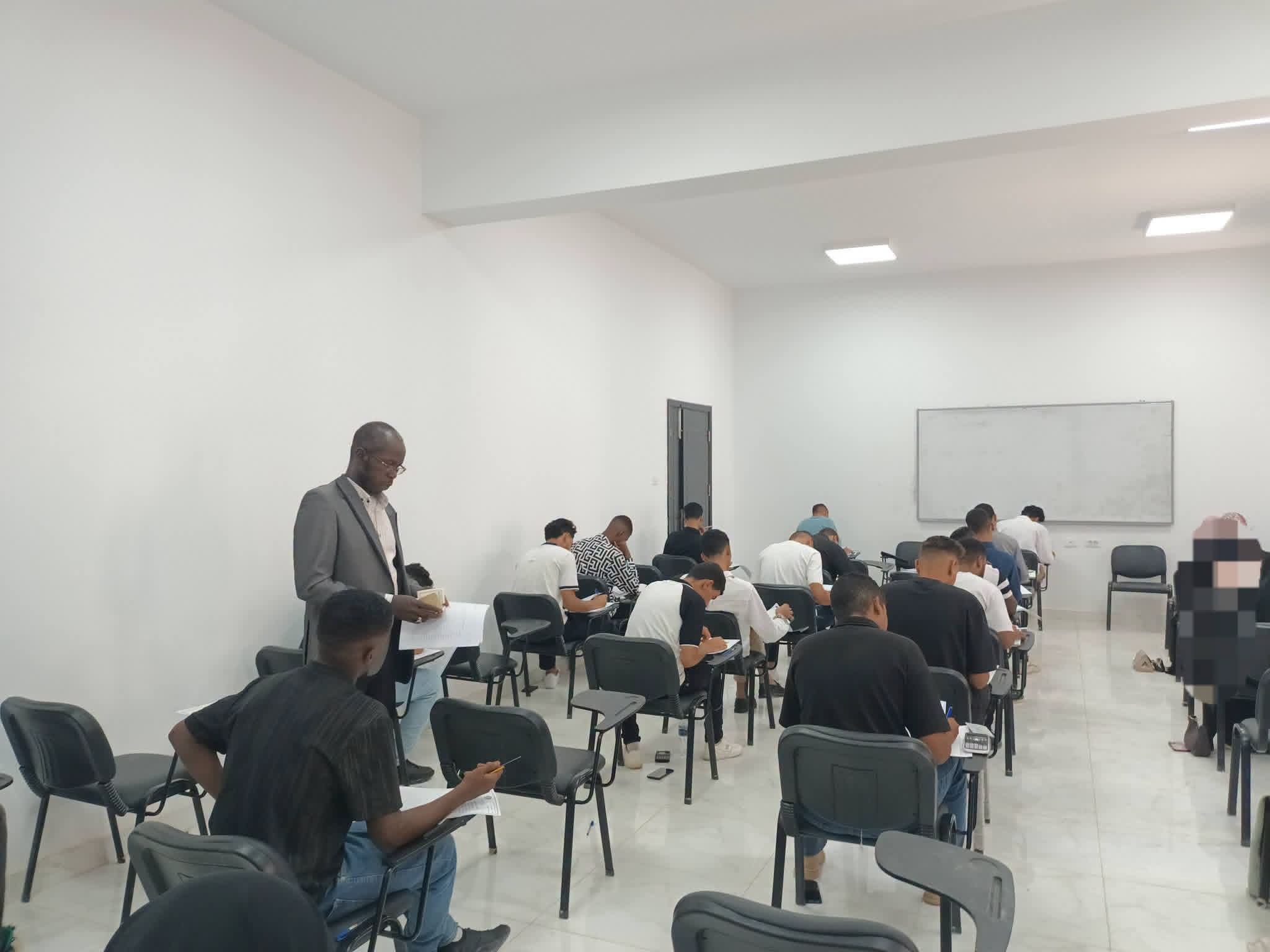Instructor in a gray suit stands at the front of a bright classroom as students write in notebooks.