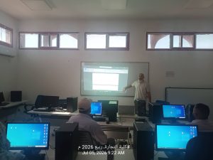 A classroom computer lab with students at desks using desktop PCs while a teacher stands at the front presenting on a projected screen; high horizontal windows line the back wall.