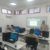Computer lab with students at desks using desktop PCs while a man stands near a projection screen at the front.