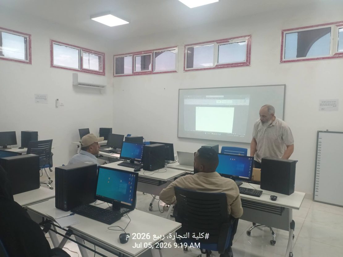 Computer lab with students at desks using desktop PCs while a man stands near a projection screen at the front.
