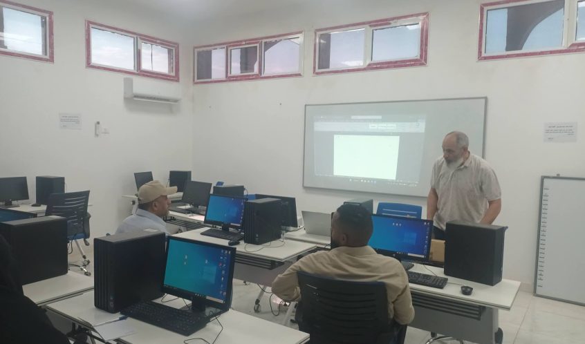 Computer lab with students at desks using desktop PCs while a man stands near a projection screen at the front.