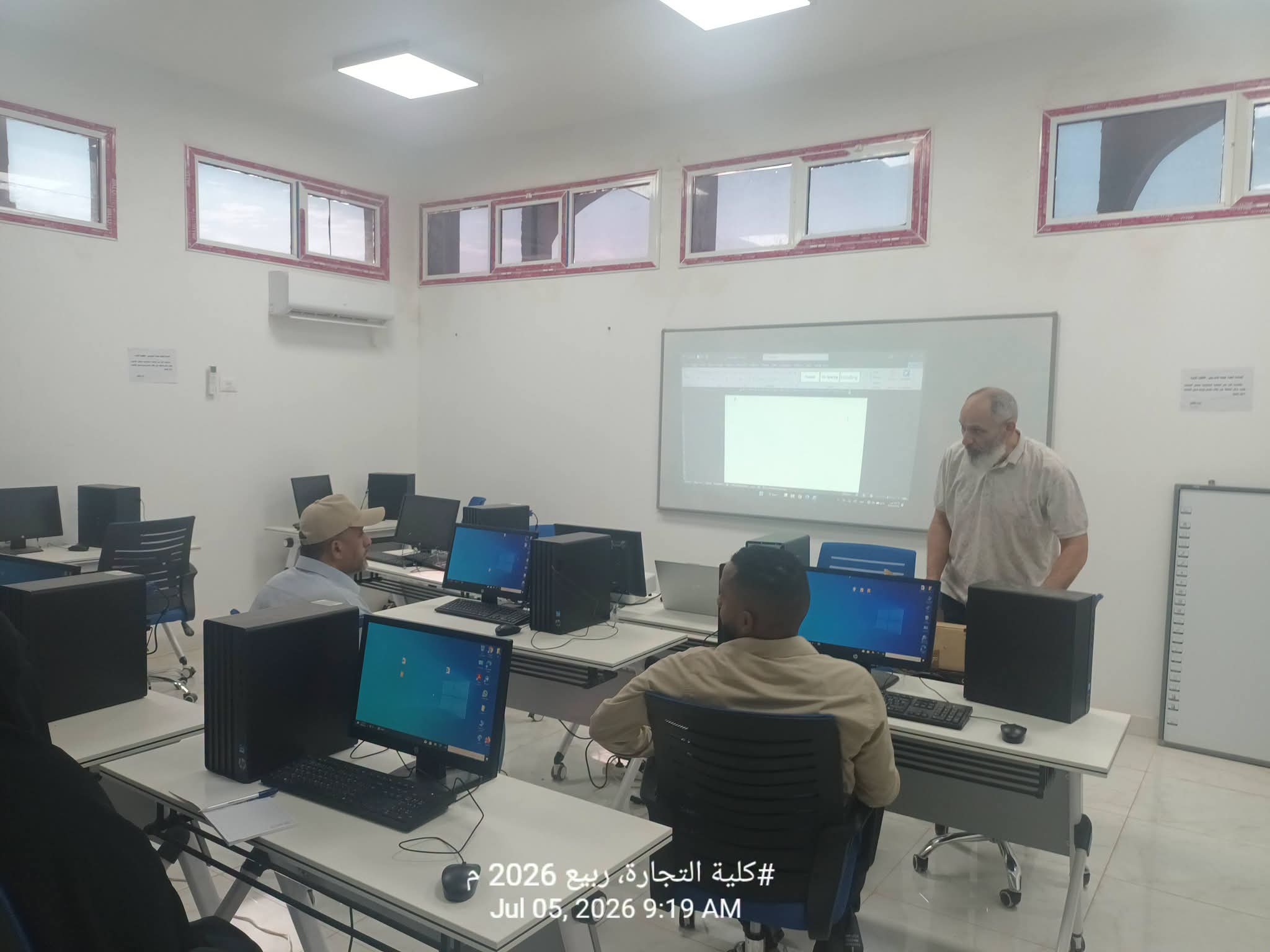 Computer lab with students at desks using desktop PCs while a man stands near a projection screen at the front.