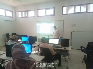 A classroom with students at computer desks facing a projection screen where a instructor stands near the board.