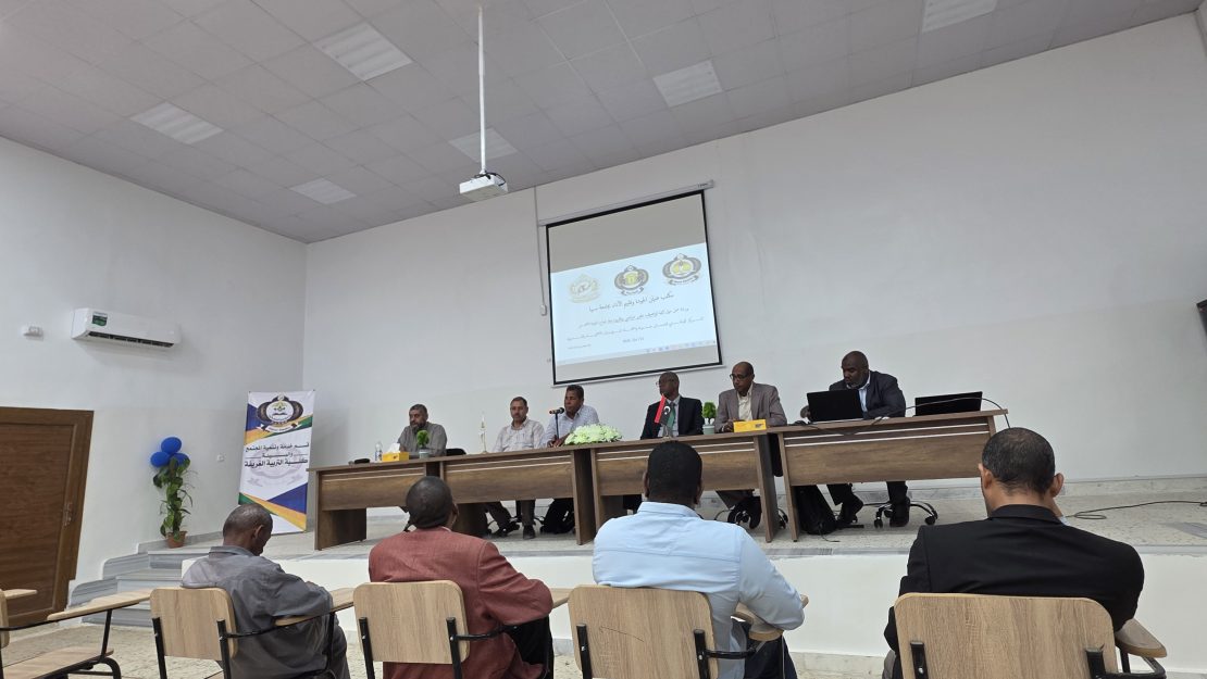 Panel of speakers seated at a long desk on a stage in a conference room, projection screen displaying a slide behind them, audience facing forward.
