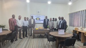 Group of men in a classroom posing for a photo in front of a mounted screen with Arabic text and logo.