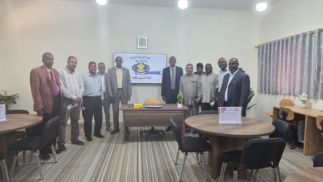 Group of about a dozen men in formal attire posing for a photo in a training room, with a screen displaying Arabic text and a logo behind them.