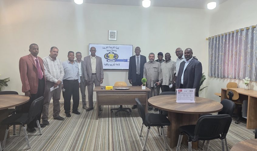 Group of about a dozen men in formal attire posing for a photo in a training room, with a screen displaying Arabic text and a logo behind them.