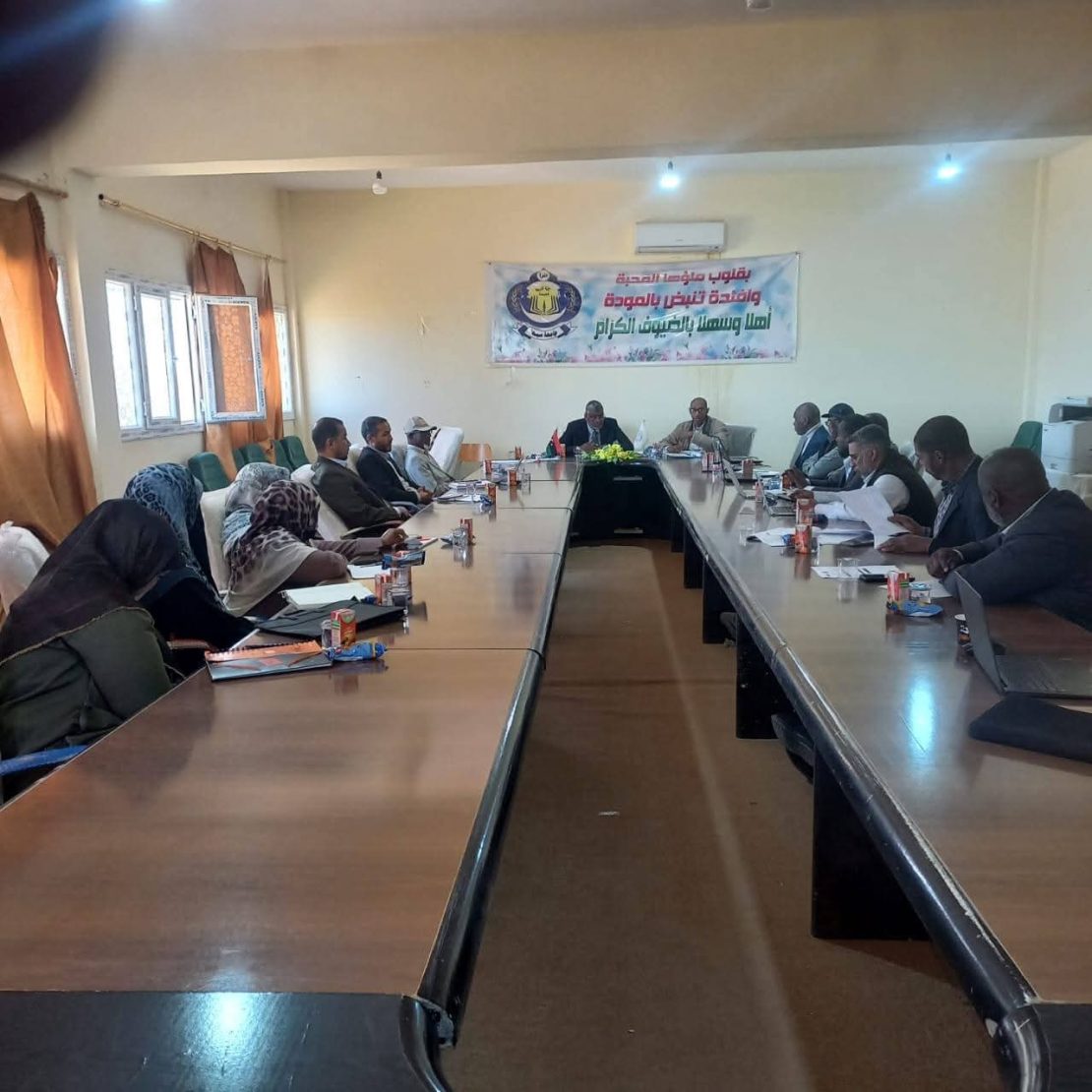A long conference table with people seated around it in a meeting room, a banner on the wall behind them with Arabic text.