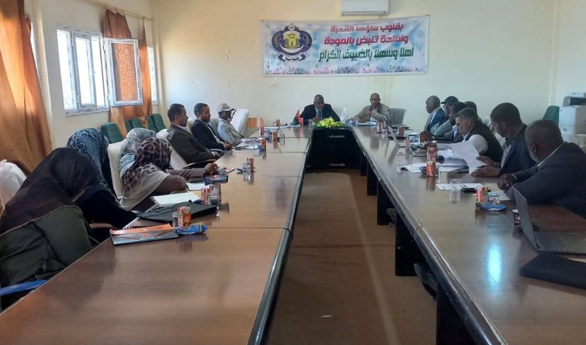 A long conference table with people seated around it in a meeting room, a banner on the wall behind them with Arabic text.