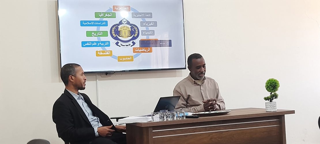 Two men sit at a wooden desk in a classroom as a slide showing Arabic school subjects around an emblem is projected on a wall monitor, with a small potted plant on the desk.