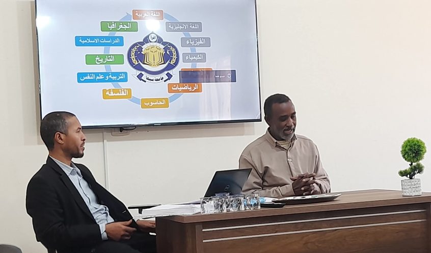 Two men sit at a wooden desk in a classroom as a slide showing Arabic school subjects around an emblem is projected on a wall monitor, with a small potted plant on the desk.