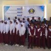 Group of students in maroon uniforms with white shirts and hijabs posing for a class photo indoors in front of a banner with a school crest and Arabic text, likely at an event.