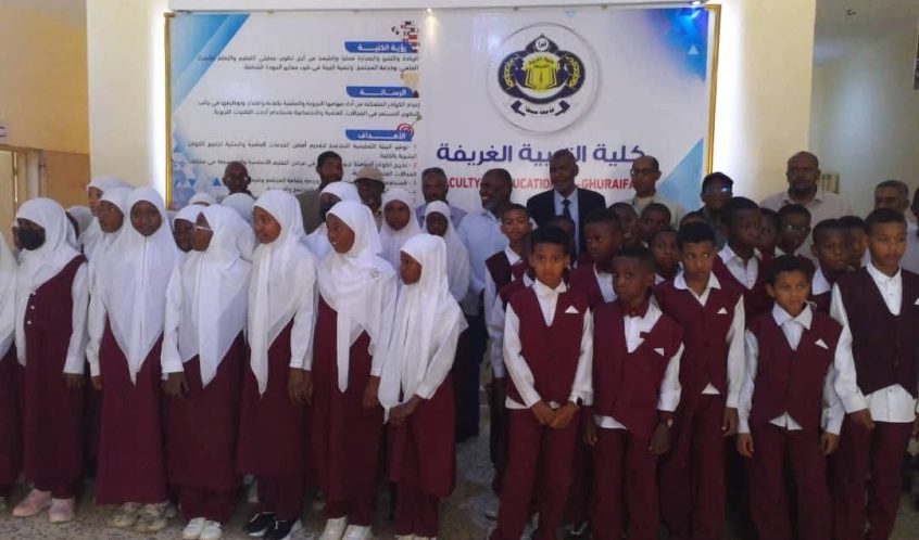 Group of students in maroon uniforms with white shirts and hijabs posing for a class photo indoors in front of a banner with a school crest and Arabic text, likely at an event.