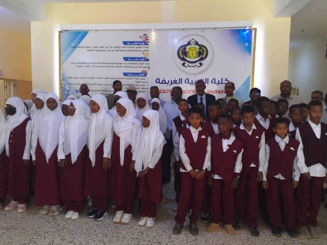 Group of students in maroon uniforms with white shirts and hijabs posing for a class photo indoors in front of a banner with a school crest and Arabic text, likely at an event.