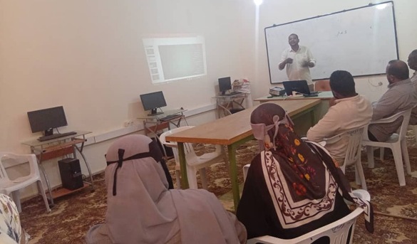 Instructor lecturing at a whiteboard in a computer-class room; adults sit at desks with desktop computers and a projection screen visible in the background.