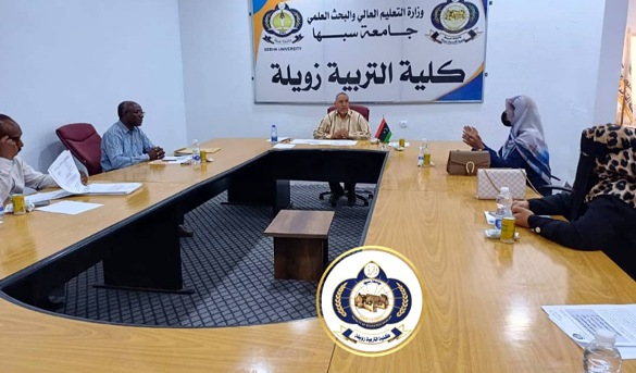 Meeting in progress in a university conference room: faculty members sit around a U-shaped wooden table, with a man at the head, a banner in Arabic on the wall, and an emblem on the foreground table edge.