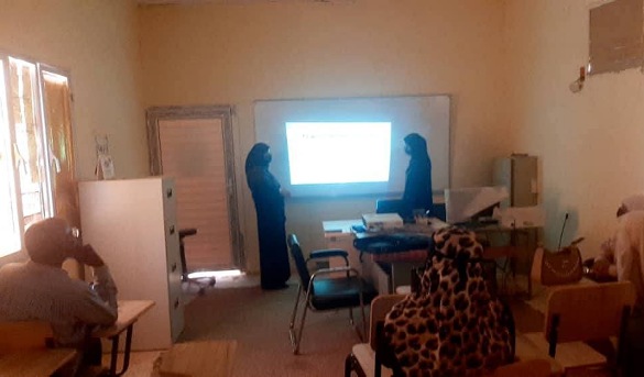 Small training room with a projector screen at the front; two presenters stand by the desk while attendees sit at desks in rows.