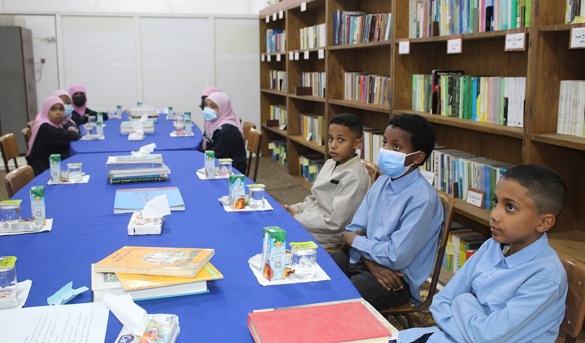 Group study session in a library: children and adults wearing masks sit along a long blue table with books and drinks.