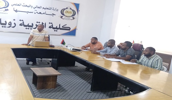 Presenter at the head of a U-shaped conference table gives a talk to five men in a university meeting room, with a large Arabic banner and small flags on the table behind them.
