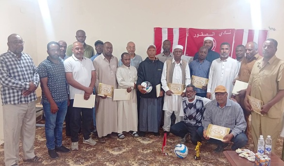 Group of men posing with certificates at a ceremony; center person holds a ball, red-and-white banner in background.
