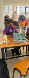 Person in a purple shirt studies at a table with an open laptop showing a colorful display; a water bottle sits nearby.