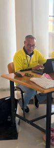 Man in a yellow polo shirt and glasses sits at a wooden table, typing on a laptop in a bright room with tall columns nearby.