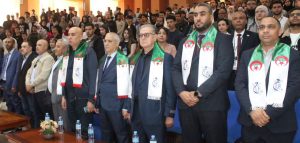 Group of men in suits wearing white scarves with Algerian flags standing at a formal ceremony, with seated audience behind them.