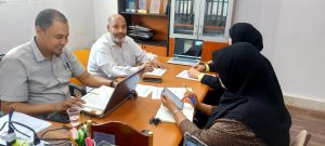 Four colleagues sit around a wooden conference table in an office, reviewing documents with laptops, notebooks, and a glass of water visible. In the photo, two men on the left and two women in hijabs on the right participate in the meeting.