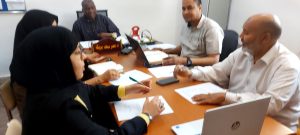 Six professionals around a conference table reviewing documents; two women in hijabs and four men, laptops and papers visible.