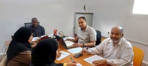 Four adults sit around a wooden conference table in a bright room, two women wearing hijabs opposite two men, with laptops, papers, and pens visible.
