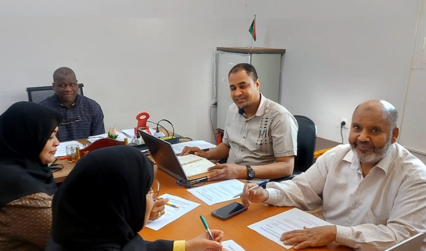 Four adults sit around a wooden conference table in a bright room, two women wearing hijabs opposite two men, with laptops, papers, and pens visible.