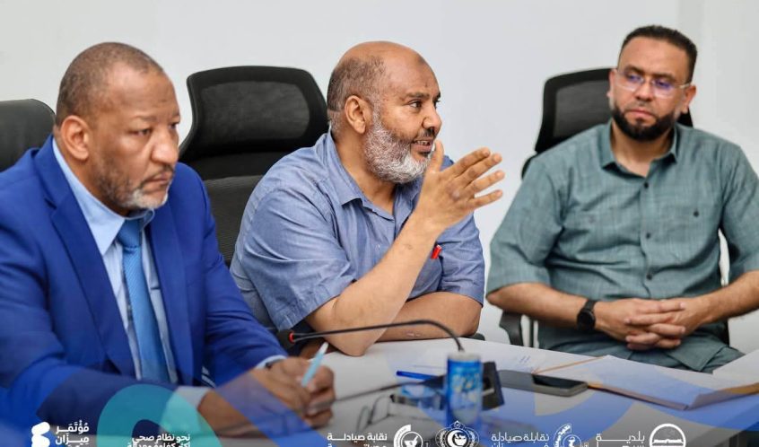 Three men in a meeting around a table; the middle man speaks with a hand gesture as the others listen, with organization logos along the bottom.