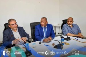 FB_IMG_1781127177819 - كلية الصيدلة - جامعة سبها Three men in a panel-style meeting seated at a conference table with microphones, papers, and drinks, the center man wearing a blue suit looking to his left.